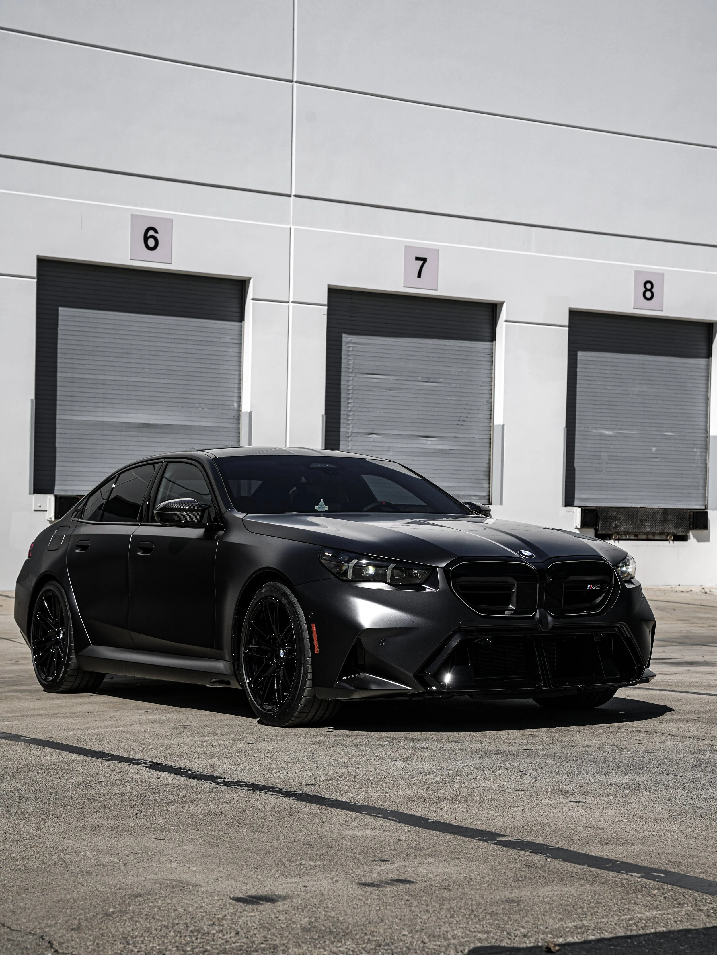A black sports car parked in front of a white building with three loading docks labeled 6, 7, and 8.