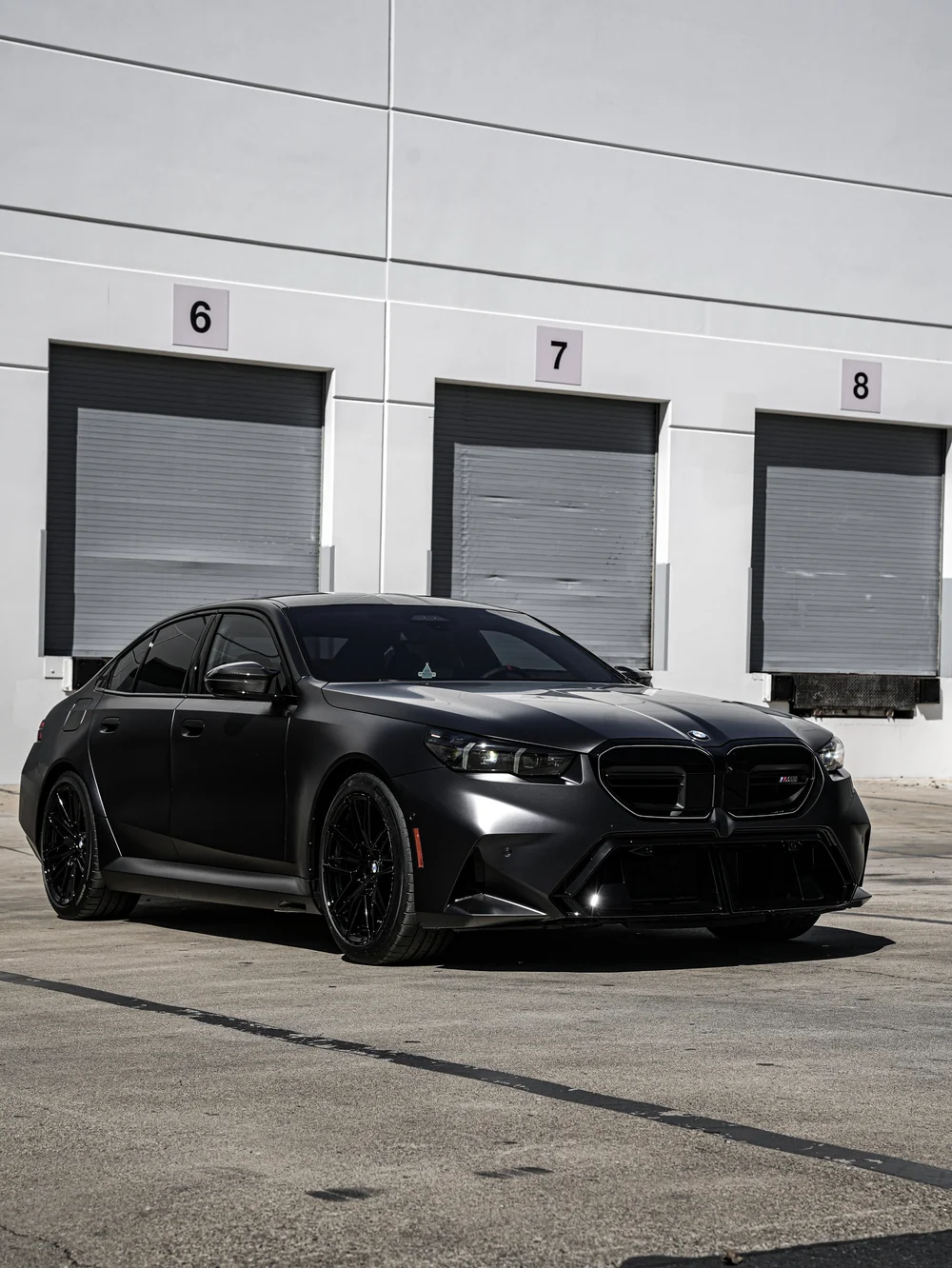BMW G90 M5 in a front three-quarter view outside numbered garage doors, with its matte hood and front grille visible