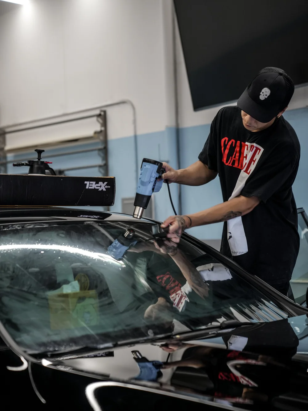 Technician holding a blue heat gun above film on the exterior of the Mercedes-AMG GLC 63 S windshield while using a small hand tool against the glass inside the shop.