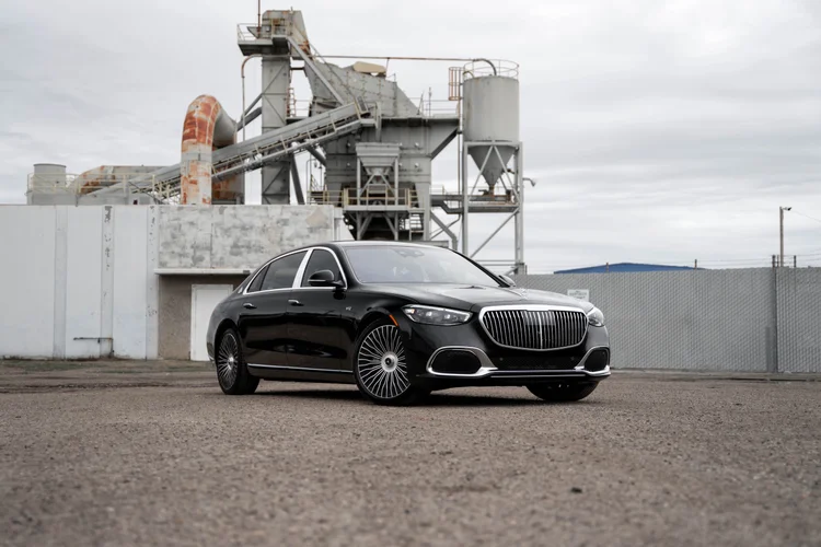 Complete front-three-quarter landscape of the black Mercedes-Maybach S 680 with a hood ornament and bright vertical grille below industrial structures
