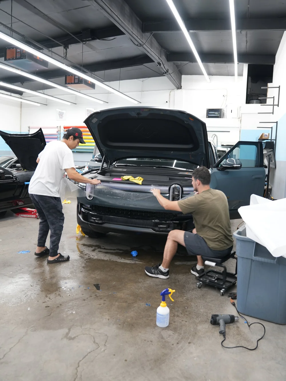 Two technicians holding a broad wet film sheet across the Rivian R1T front body panel below its light bar, with the hood and a front door open in the studio.