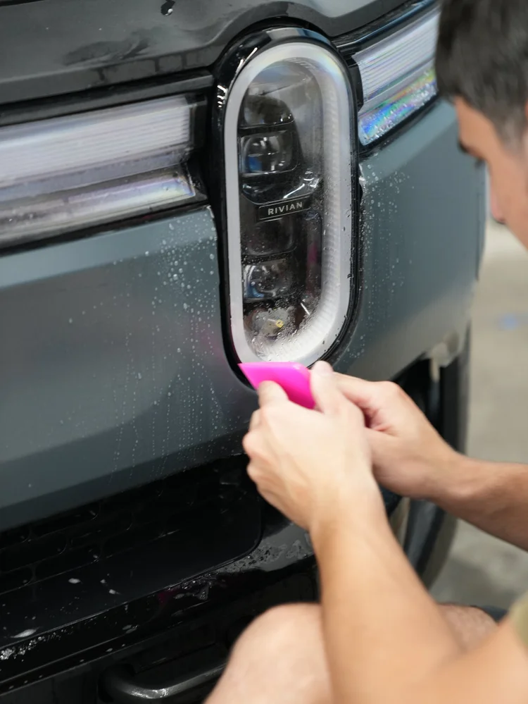 A technician holding a pink squeegee beside wet film along the lower edge of a Rivian R1T oval headlamp.