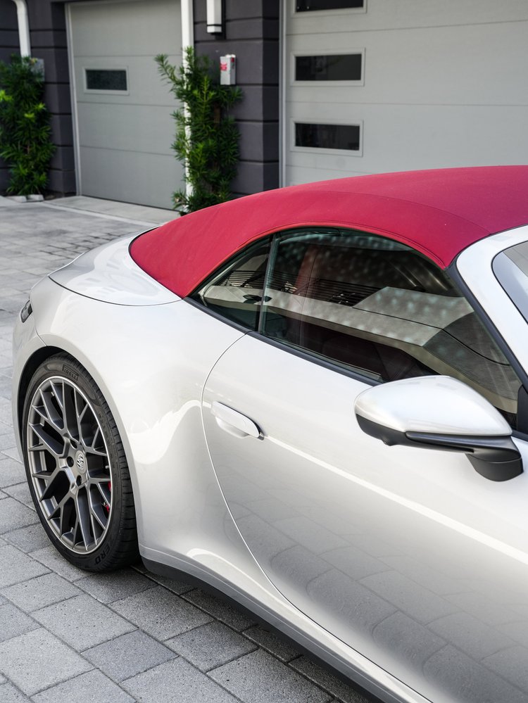 Side detail of the silver Porsche 911 with its red soft top raised, closed door and side glass, silver door handle and mirror, rear wheel and building reflections.