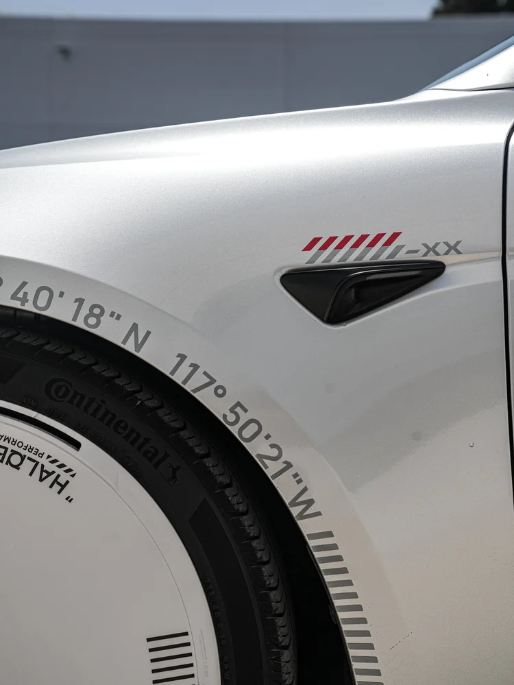Coordinate lettering and red and grey directional marks on the silver Model 3 front fender beside its side camera and partial wheel cover.