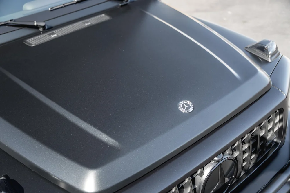 High hood detail of the grey G 63 showing matte-looking contours, a round Mercedes badge, wipers and the grille below