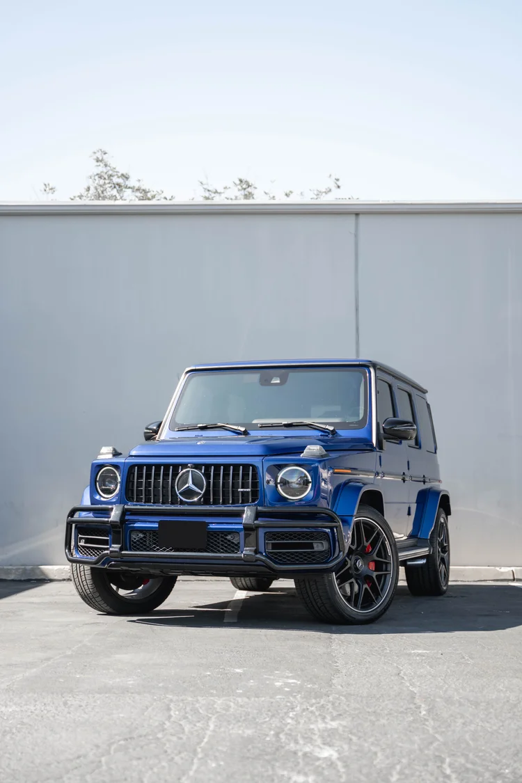 Tall complete front-three-quarter view of the blue G 63 with its circular lamps and vertical grille below a white wall