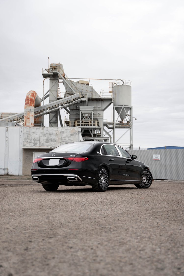Tall complete rear-three-quarter portrait of the black Maybach beneath a cloudy sky and industrial towers