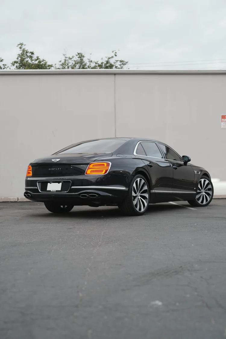 Rear three-quarter portrait of the dark Bentley against a pale wall.