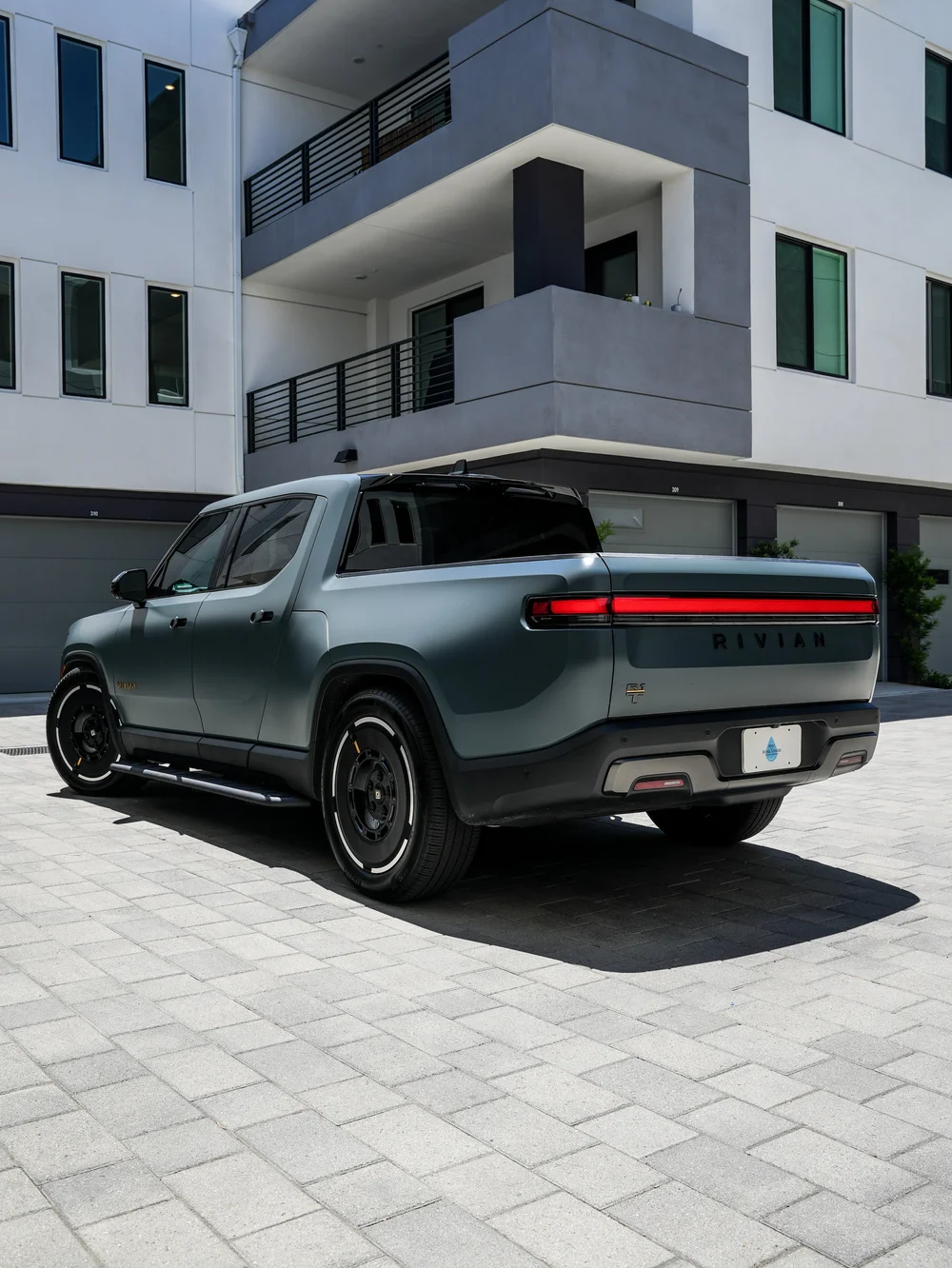 Complete left-facing rear three-quarter view of the satin-finish Rivian R1T with dark glass, black wheels, red rear light bar and side step on courtyard paving.
