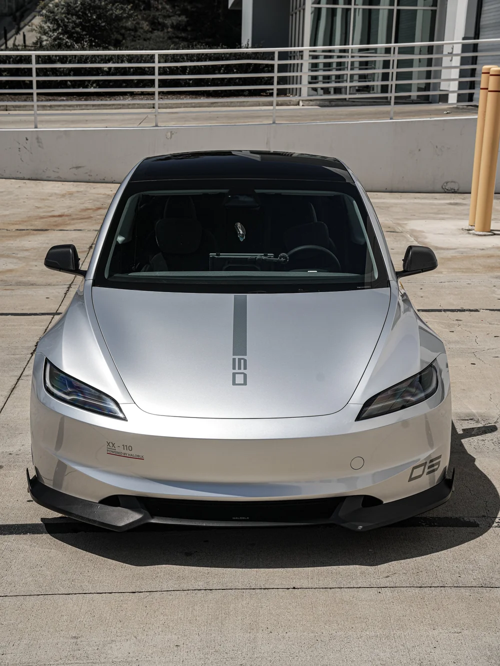 Centered front view of the silver Model 3 showing hood graphics, both mirrors, headlights, front bumper and dark splitter on pavement.