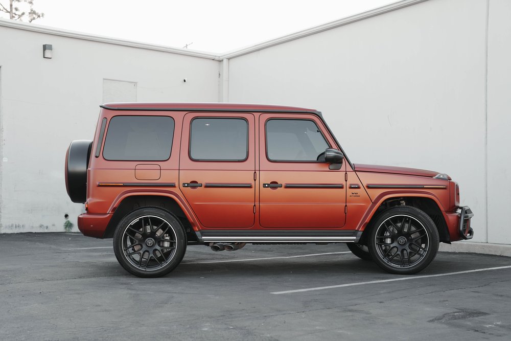Full side profile of the matte red G63 with its black wheels, dark glass, silver step and rear spare-wheel cover