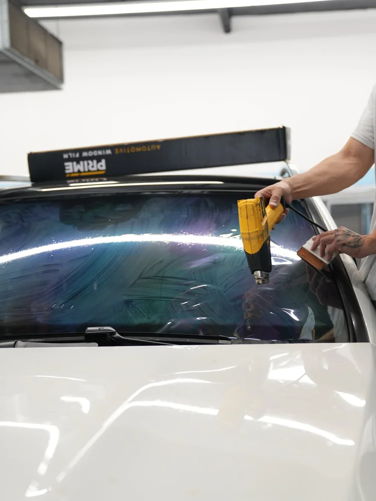 Yellow heat gun and hand tool working over wet film on the windshield exterior with a window-film box in the background.