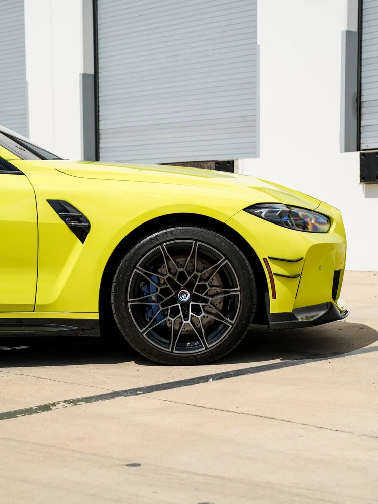 Close BMW M4 front wheel with a blue brake caliper, brake disc, side vent, part of a headlight and dark lower front trim against yellow-green bodywork.