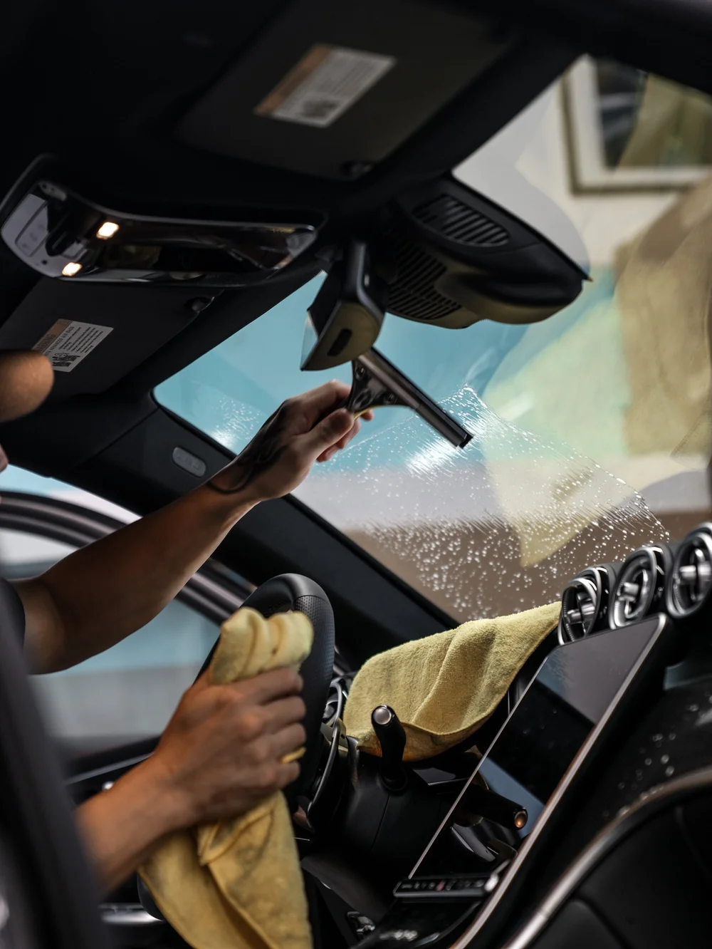 Inside the Mercedes-AMG GLC 63 S, a technician uses a wide hand squeegee against wet windshield film with yellow towels over the steering wheel and dashboard.