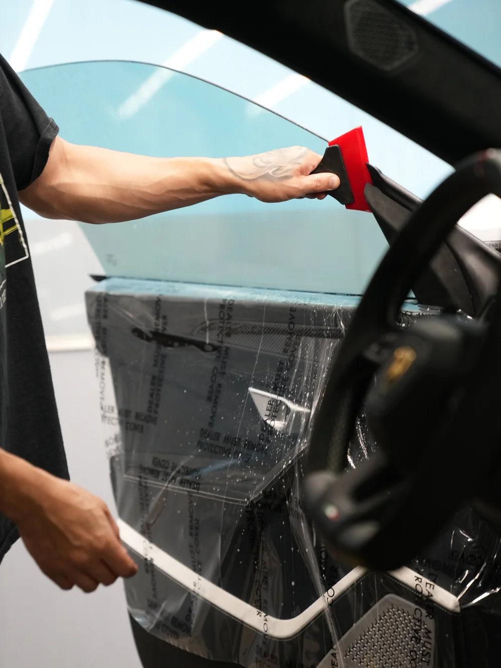 Red squeegee smoothing film on a side window above the retained door trim protected by clear plastic.