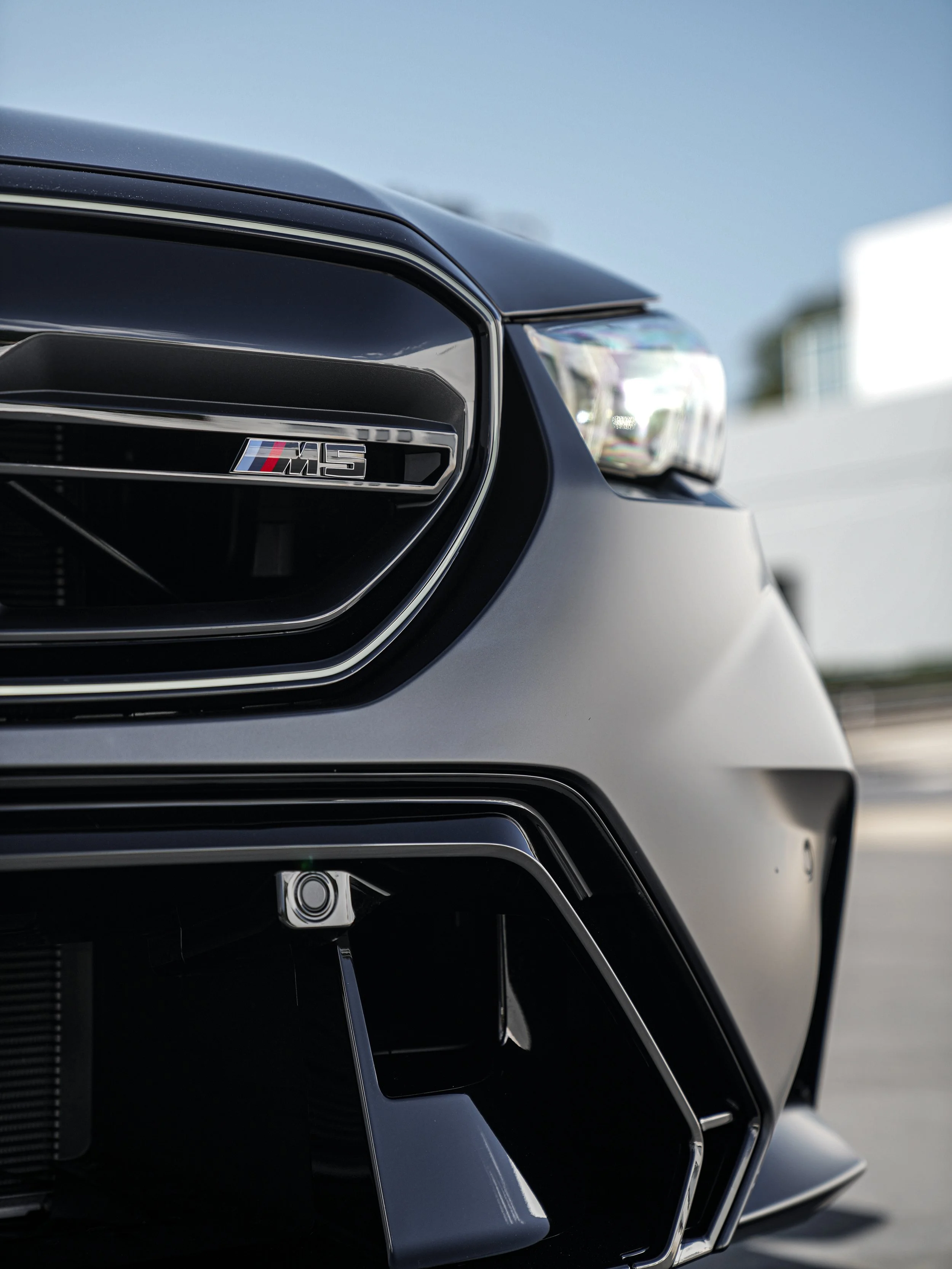 Close-up of front grille and headlight of a black Mercedes-Benz AMG vehicle, showing an M5 badge, sports car design elements, and a parking camera.