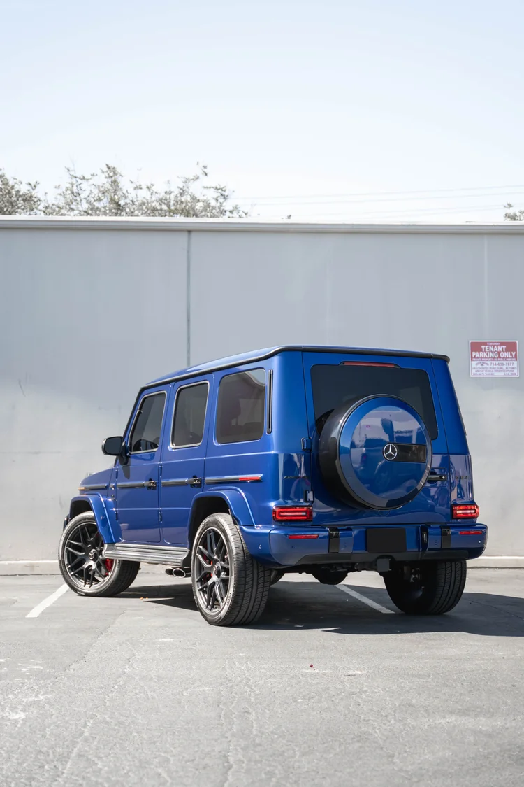 Tall complete rear-three-quarter portrait of the blue G 63 below a white wall and pale sky