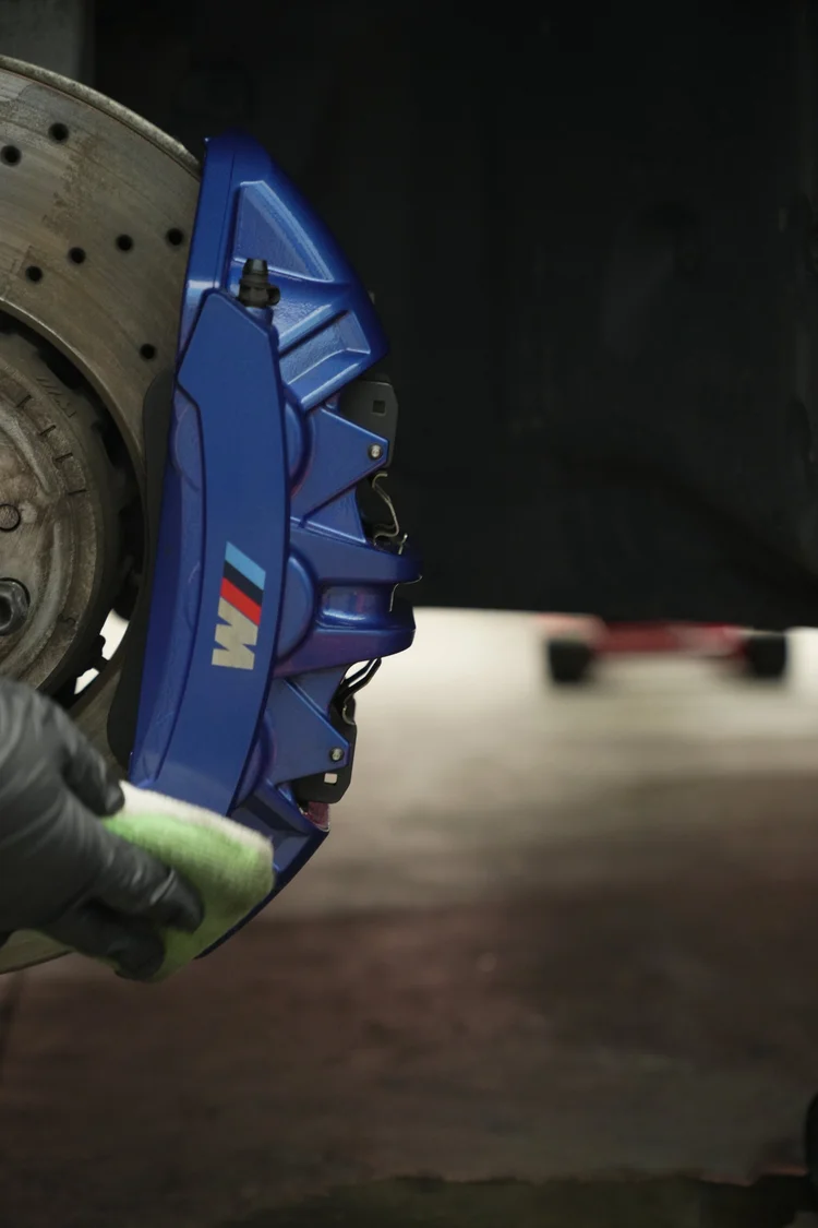 Gloved hand holding a white and green applicator against a blue M-lettered caliper beside a drilled brake rotor.