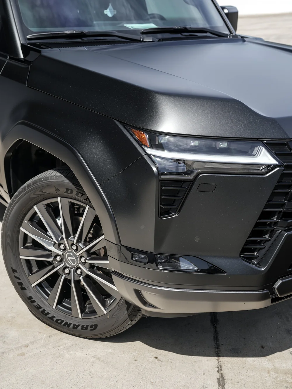 Close Lexus front corner with a dark-and-bright multispoke wheel, hood ridge and angular illuminated headlamp beside the cropped grille