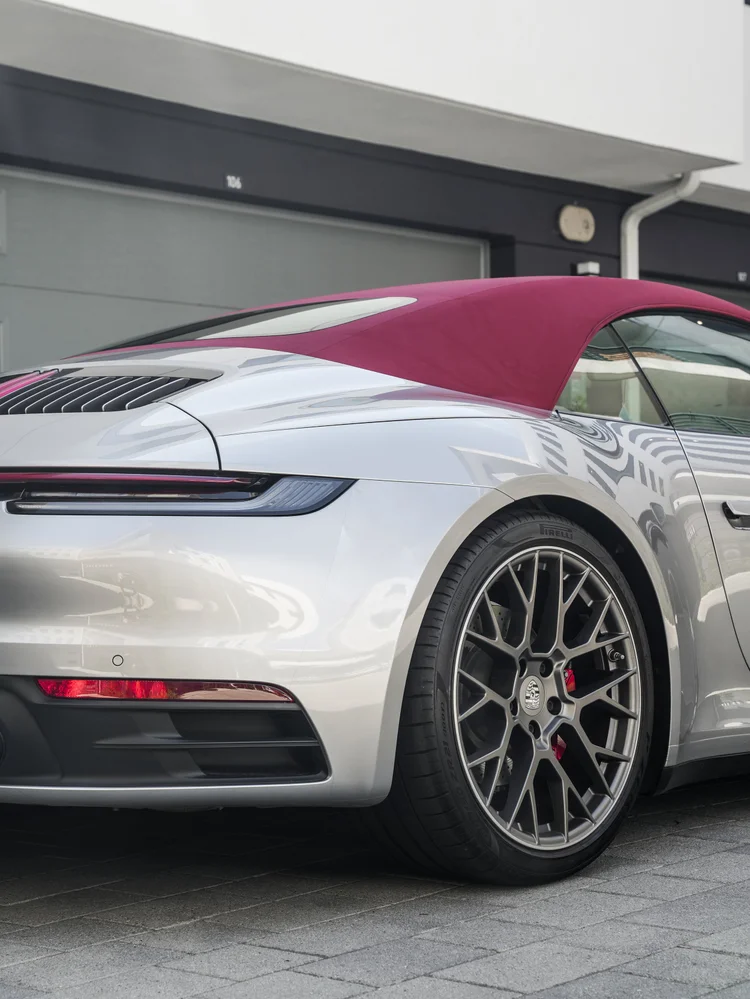 Rear-quarter detail of the silver Porsche 911 with its red soft top raised, rear lamp strip, dark multi-spoke wheel, red brake caliper and building reflections.