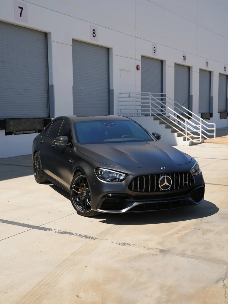 Complete front three-quarter view of the dark matte Mercedes-AMG E 63 S with black wheels and yellow-gold brake calipers on pale pavement.