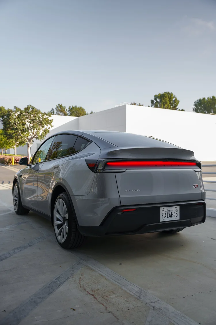 Rear three-quarter view of the gray Tesla Model Y showing the red light bar, liftgate, side glass and silver wheels with a white building and trees behind.