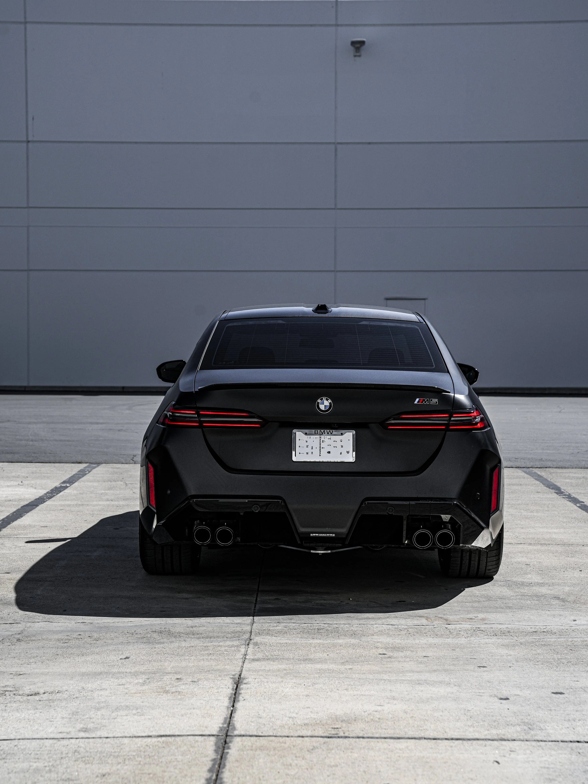 Black BMW M3 parked in an empty lot, viewed from the rear with shadow cast on the ground.