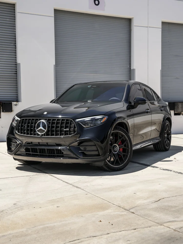 Complete front three-quarter view of the black Mercedes-AMG GLC 63 S facing left, with a vertical-bar grille, dark multi-spoke wheels, red brake calipers and dark side glass outside gray workshop doors.