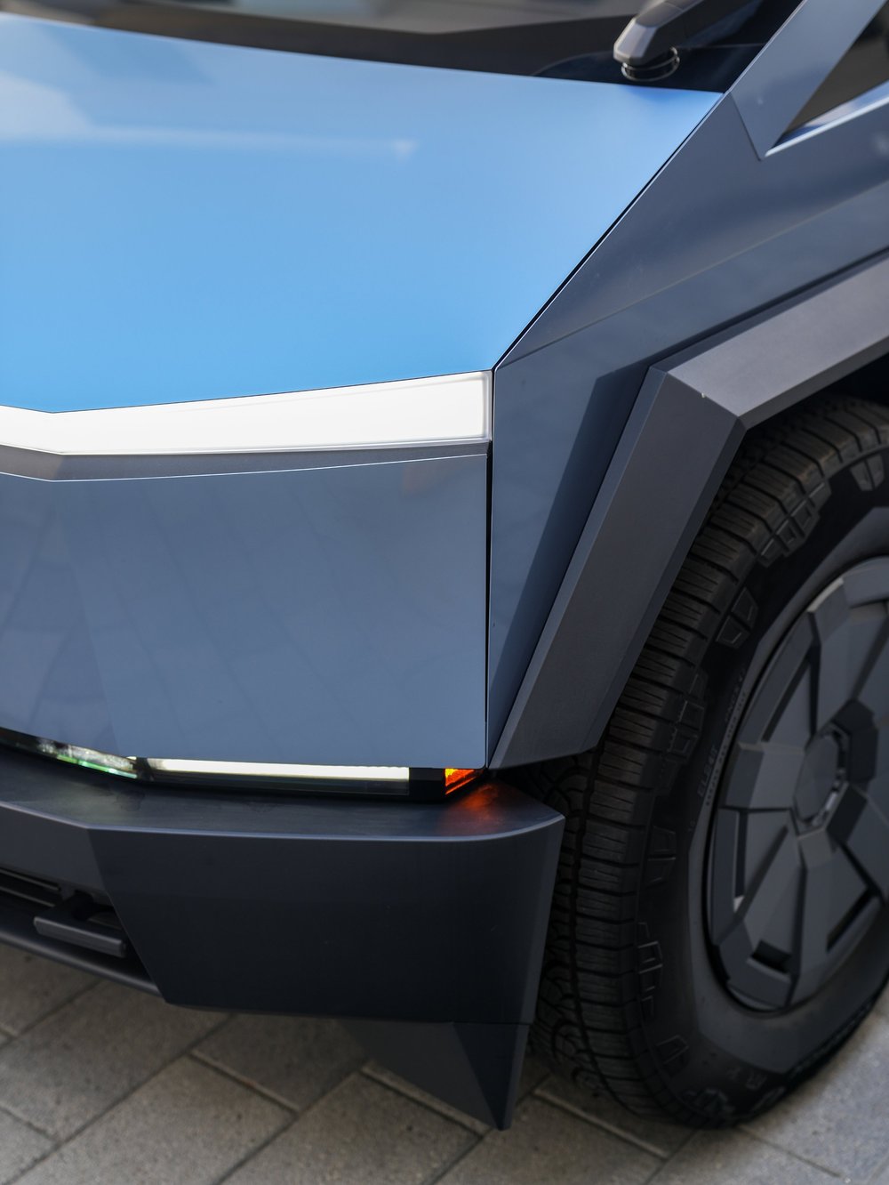 Close front corner of the Cybertruck showing the blue-gray hood and face, white lamp strip, front tire and angular black wheel-arch trim.