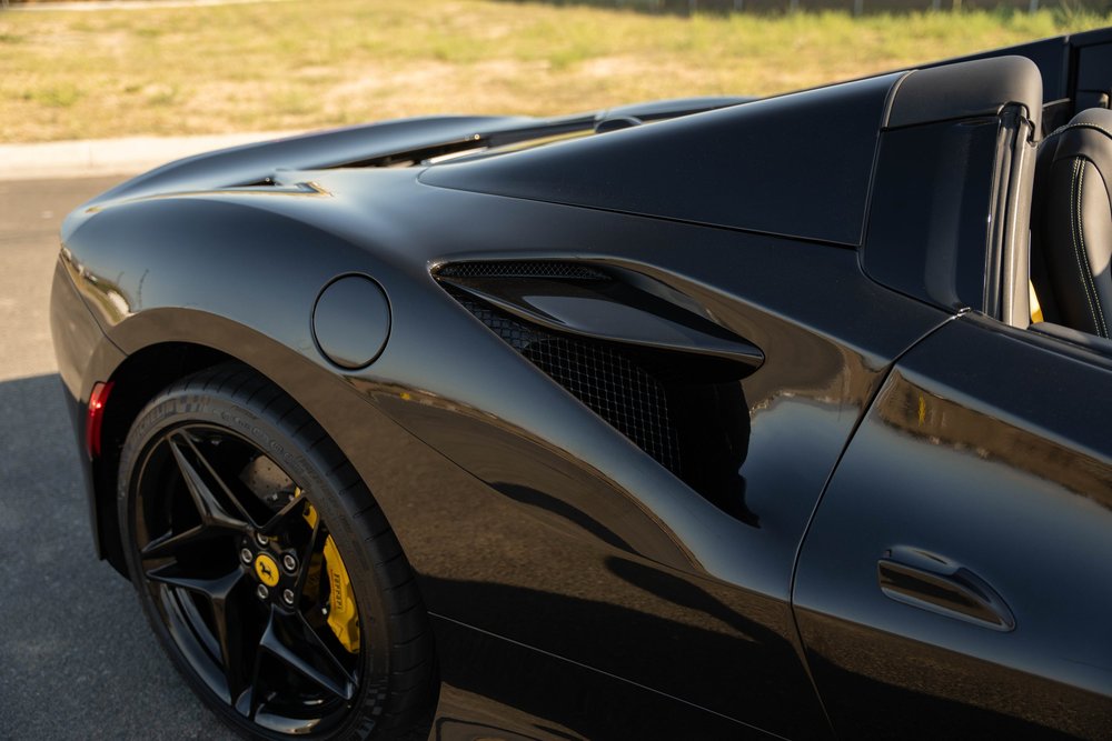 Black Ferrari F8 rear fender close-up showing side intake, fuel door, door handle and rear wheel