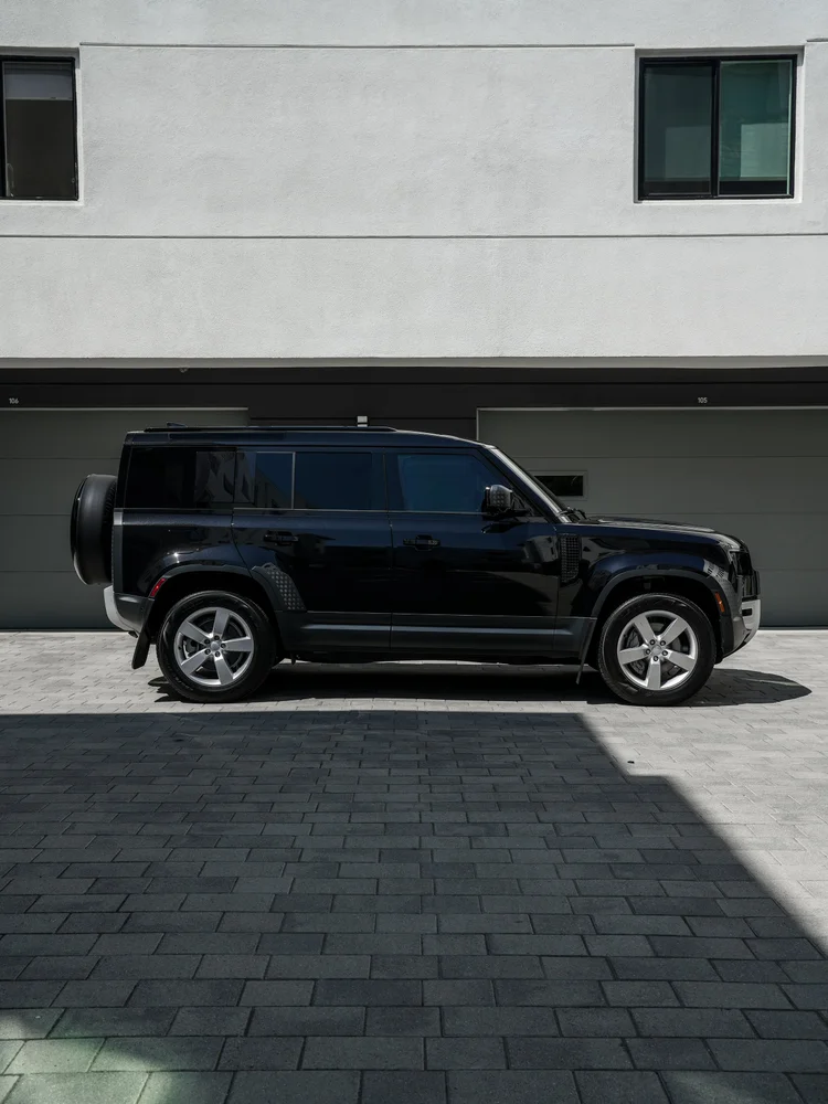 Complete side profile of the dark Defender showing silver wheels, roof rails and the rear spare-wheel cover against a pale industrial wall