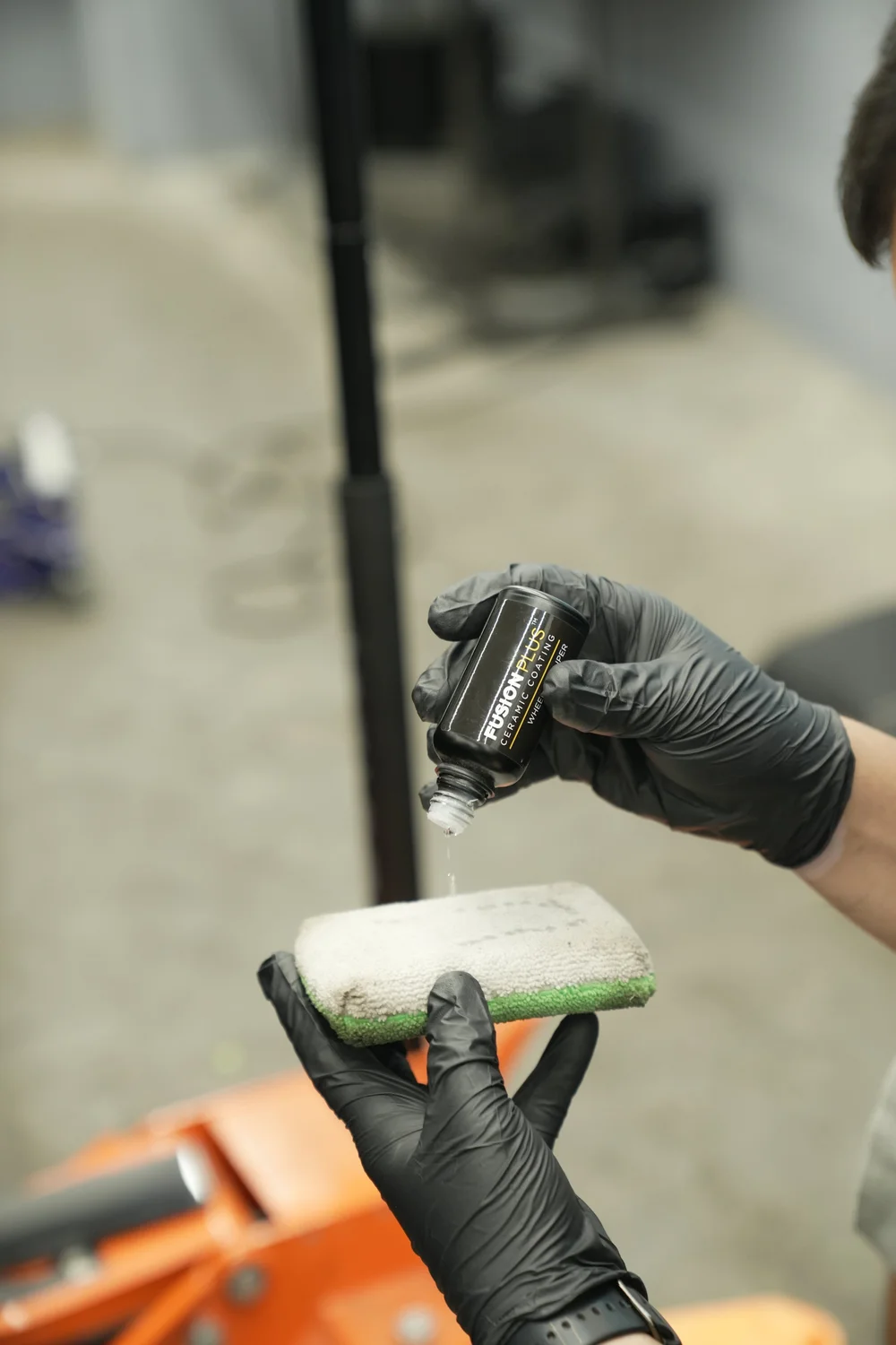 Gloved hands pouring liquid onto a white and green applicator from a bottle labeled FUSION PLUS CERAMIC COATING WHEEL & CALIPER.