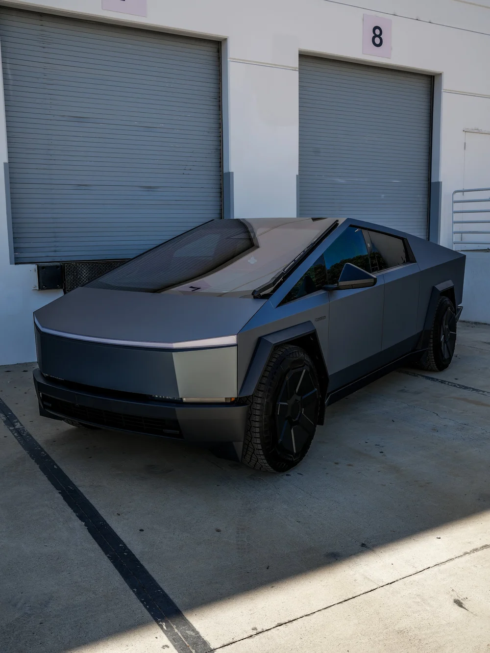 Complete left-facing front three-quarter view of the grey blue Tesla Cybertruck with dark wheels and reflective glass against workshop door eight.