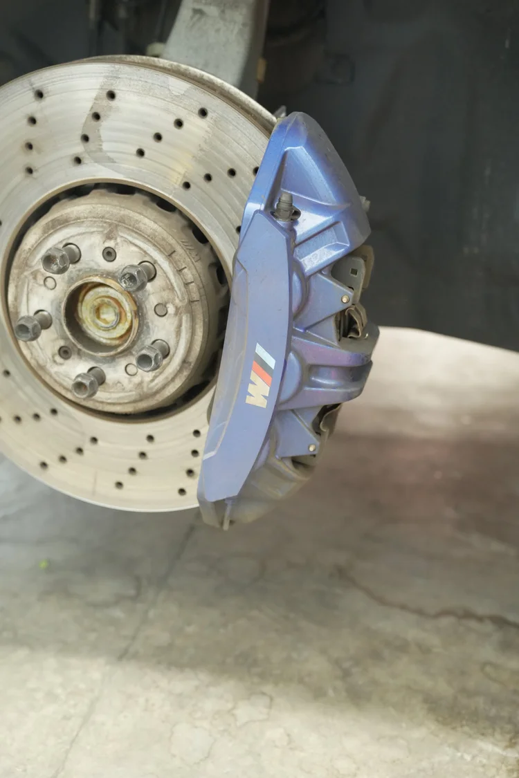 BMW M8 brake assembly with the wheel absent, showing a drilled rotor, exposed hub and blue M-lettered caliper.