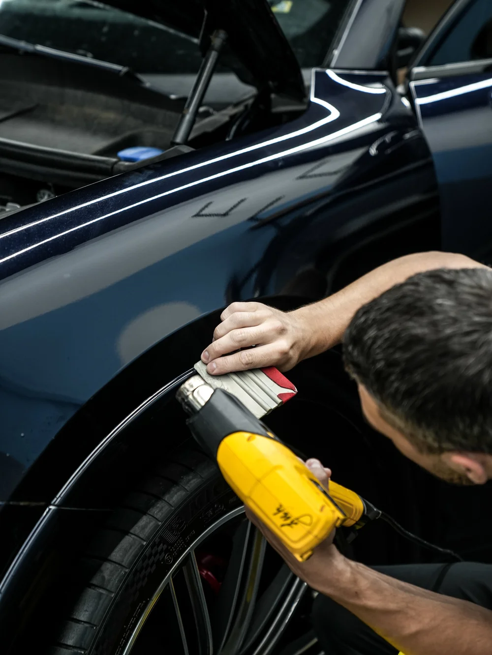 Technician holding a heat gun and padded hand tool along the Porsche Cayenne GTS front wheel-arch edge.