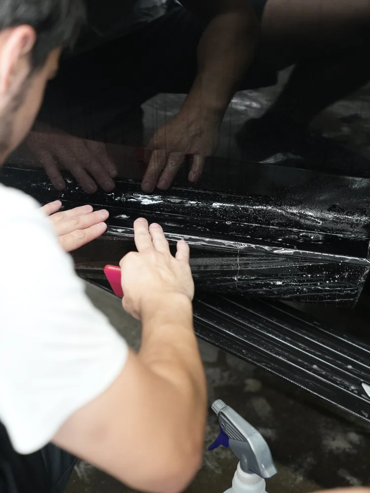 Hands positioning wet transparent film along a black side panel beneath the window trim.