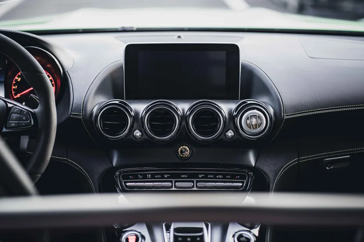 Centered dark dashboard with an unlit screen, four circular vents, yellow stitching and a yellow knob.