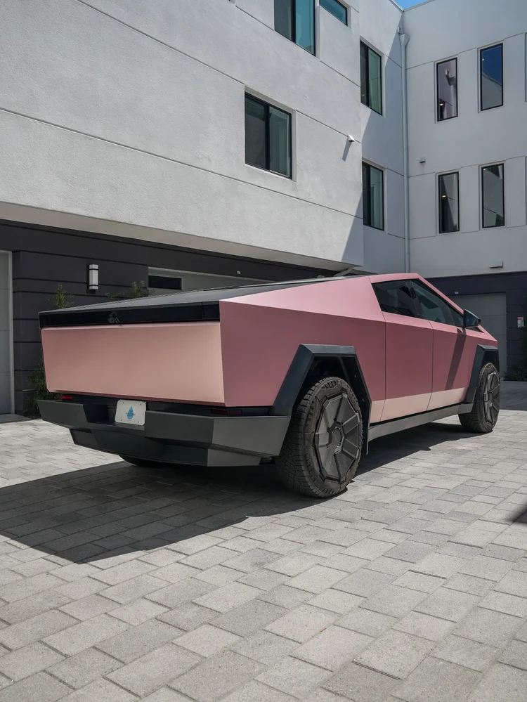 Rear three-quarter view of the pink-gold Cybertruck with its rectangular tailgate, dark bed cover, black bumper and rear wheel.