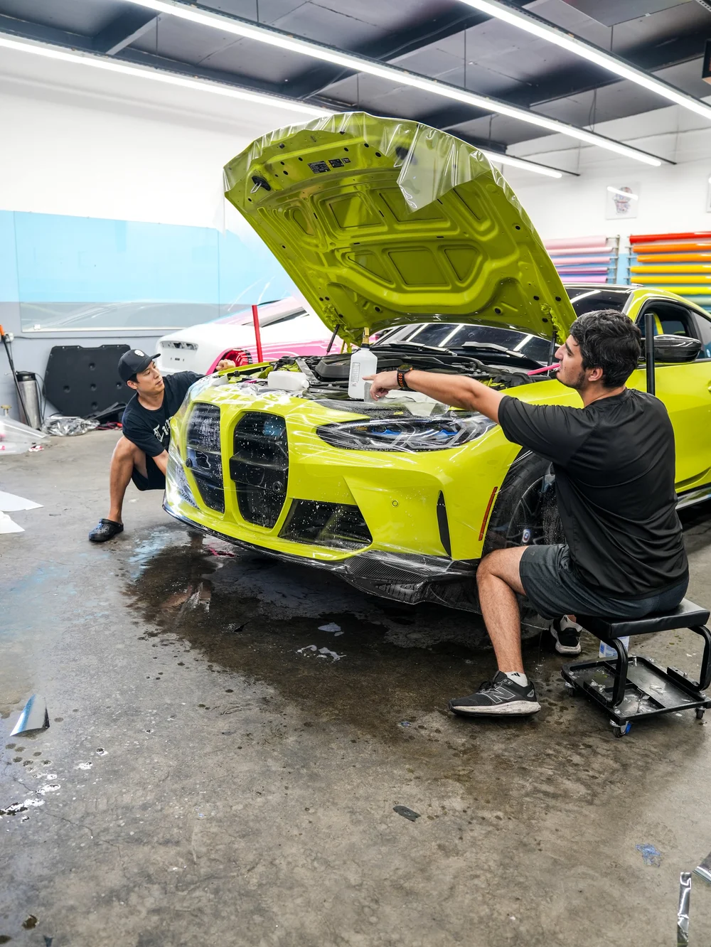 Two technicians working at the front of the yellow-green BMW M4 inside the shop, with the hood raised and wet transparent film across the front area.