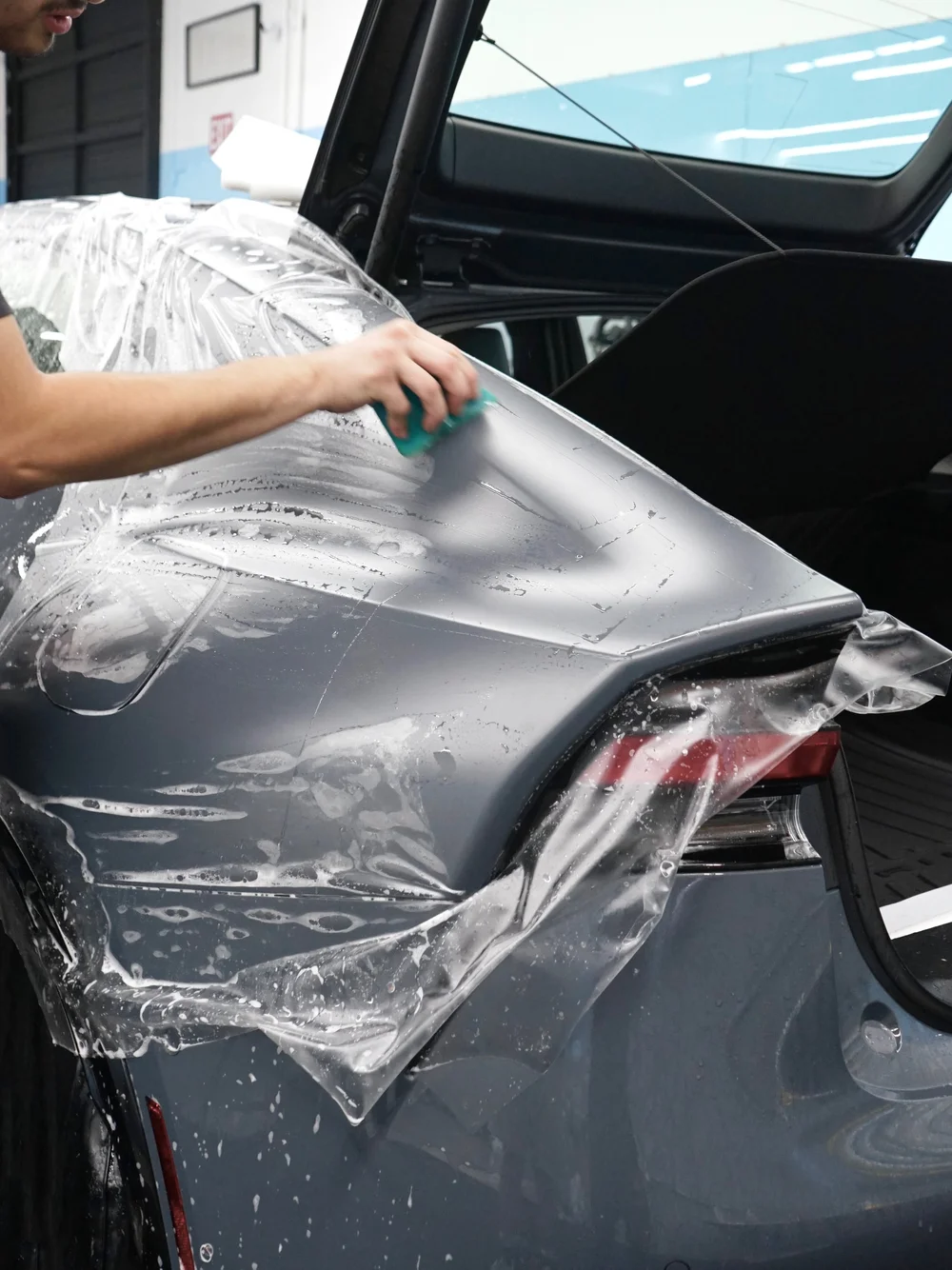 Technician fitting paint protection film to the rear panel of a gray vehicle