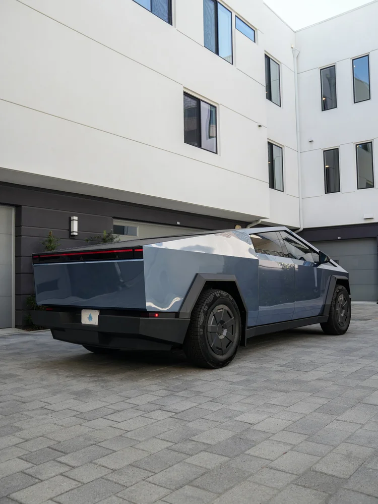 Complete rear three-quarter view of the blue-gray Cybertruck with a red rear lamp strip, reflected buildings along the side panels and dark wheels.