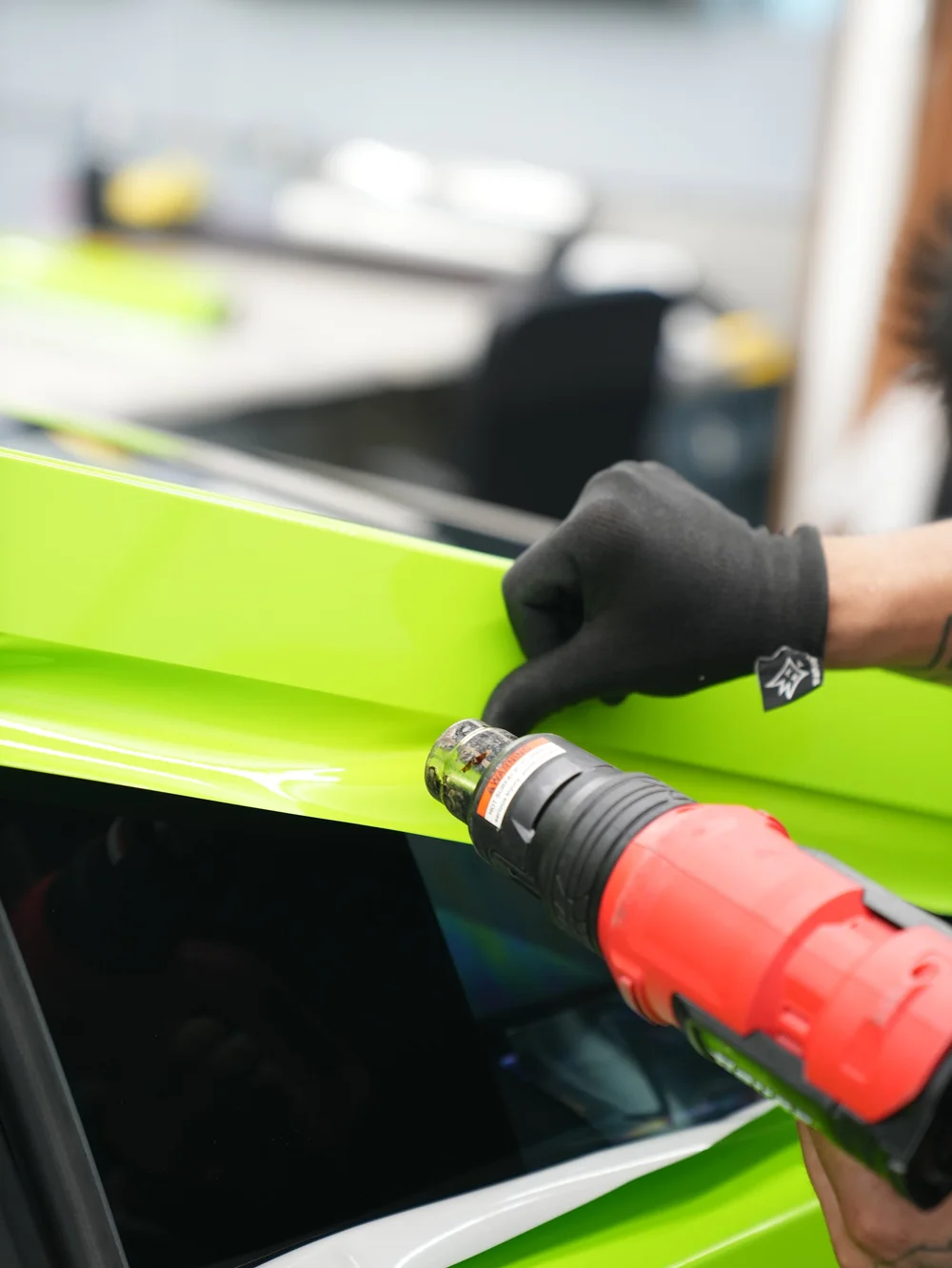 Close installation photograph showing a heat gun near glossy green vinyl and a gloved hand guiding the film along a panel edge.