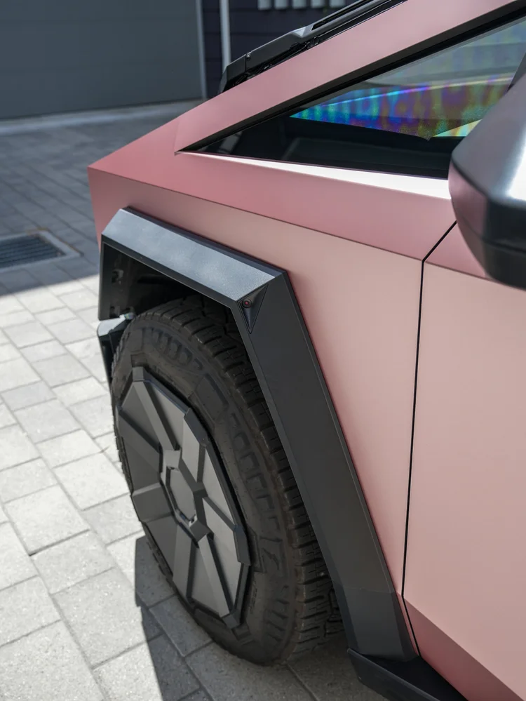 Close front wheel and fender view of the pink-gold Cybertruck with dark wheel-arch trim, mirror and reflections in the side glass.
