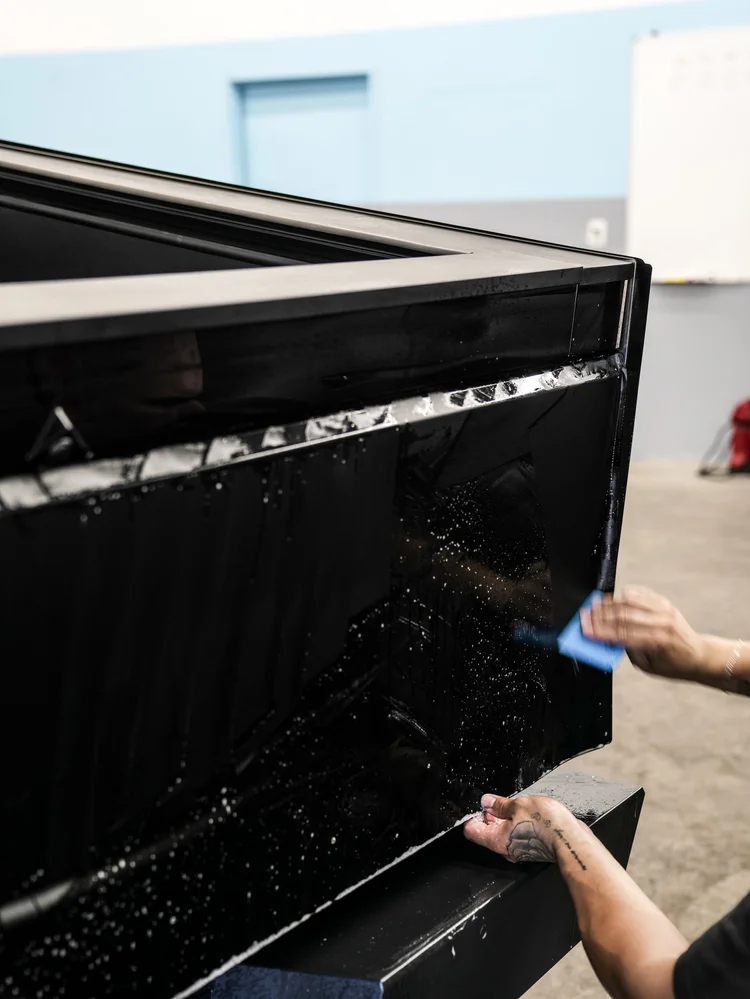 Hands holding and squeegeeing wet black film across an upright Tesla Cybertruck panel near its tailgate edge.