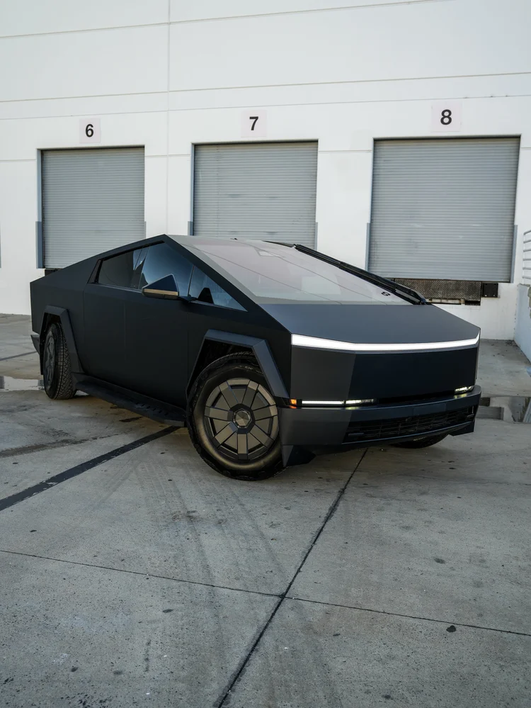Matte black Tesla Cybertruck in a front three-quarter view outside grey roller doors, with grey angular wheel covers and illuminated front light strips.