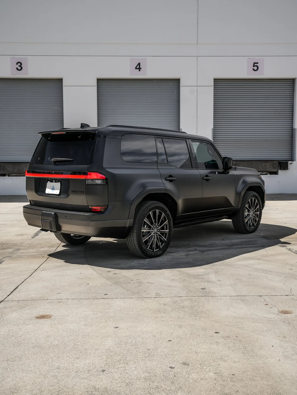 Complete rear-three-quarter view of the matte-looking GX 550 with dark side glass, bright wheel spokes and an illuminated red rear light bar on pavement