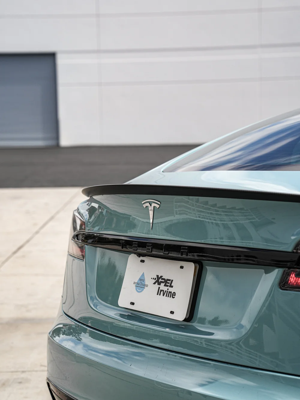 Close view of a Tesla Model S rear spoiler, Tesla emblem, rear light trim and Toyz/XPEL Irvine display plate.