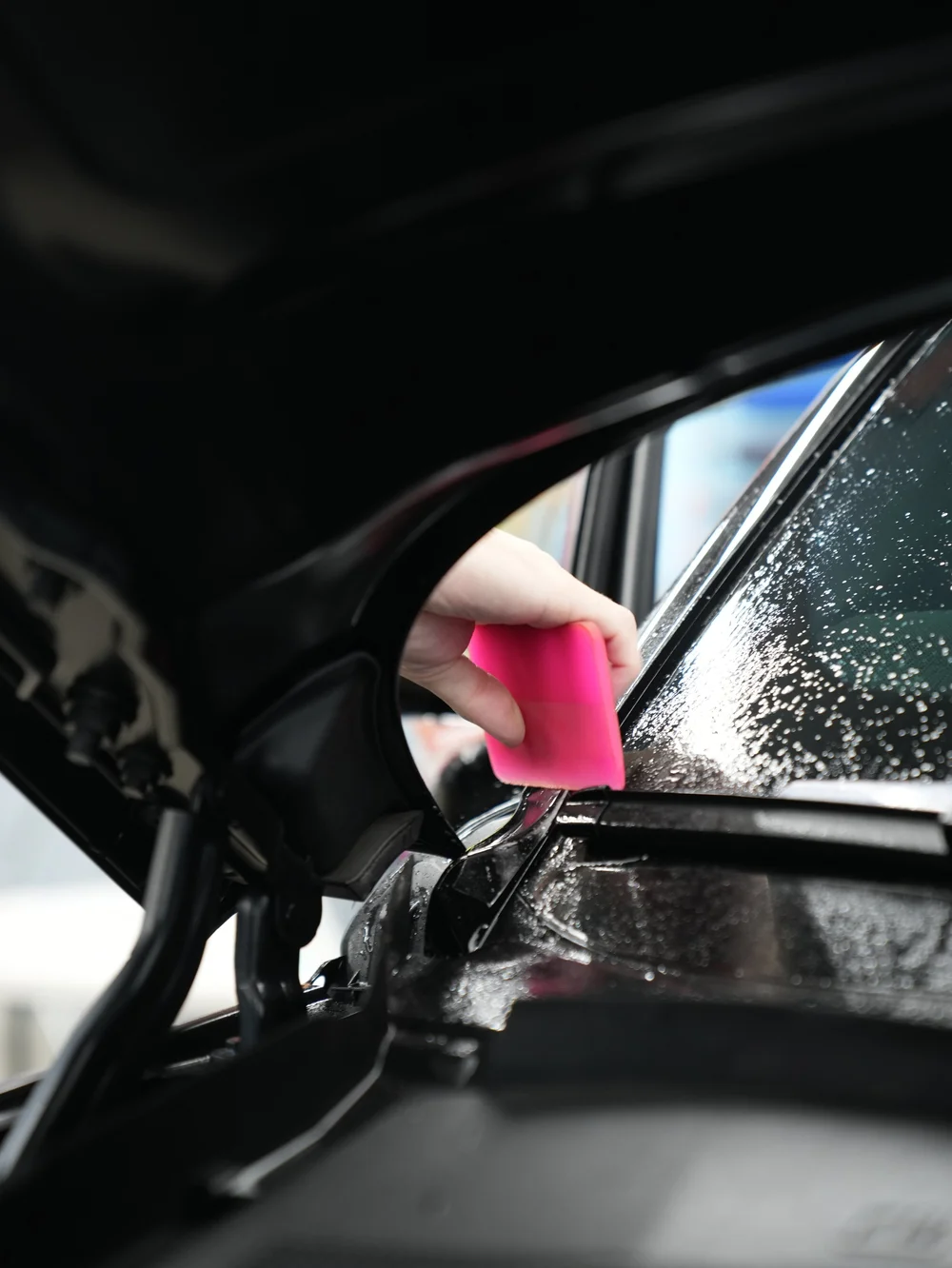 Hand using a pink squeegee along a wet glass edge beside black trim.