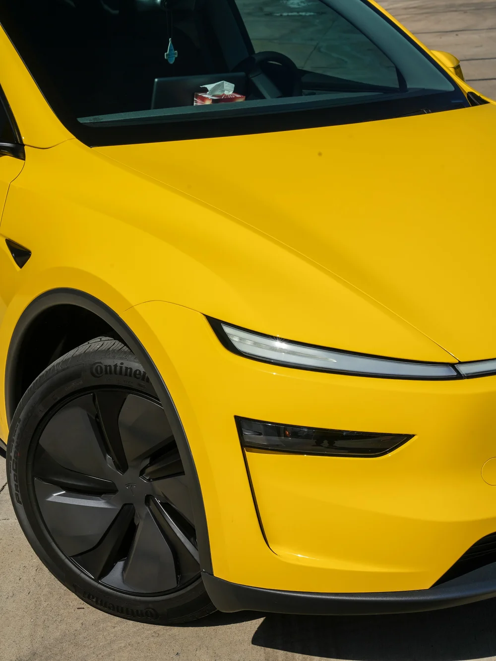 Close yellow front corner with a dark Tesla-centered wheel, white light strip and lower lamp beside the hood and windshield; the nose is cropped at right