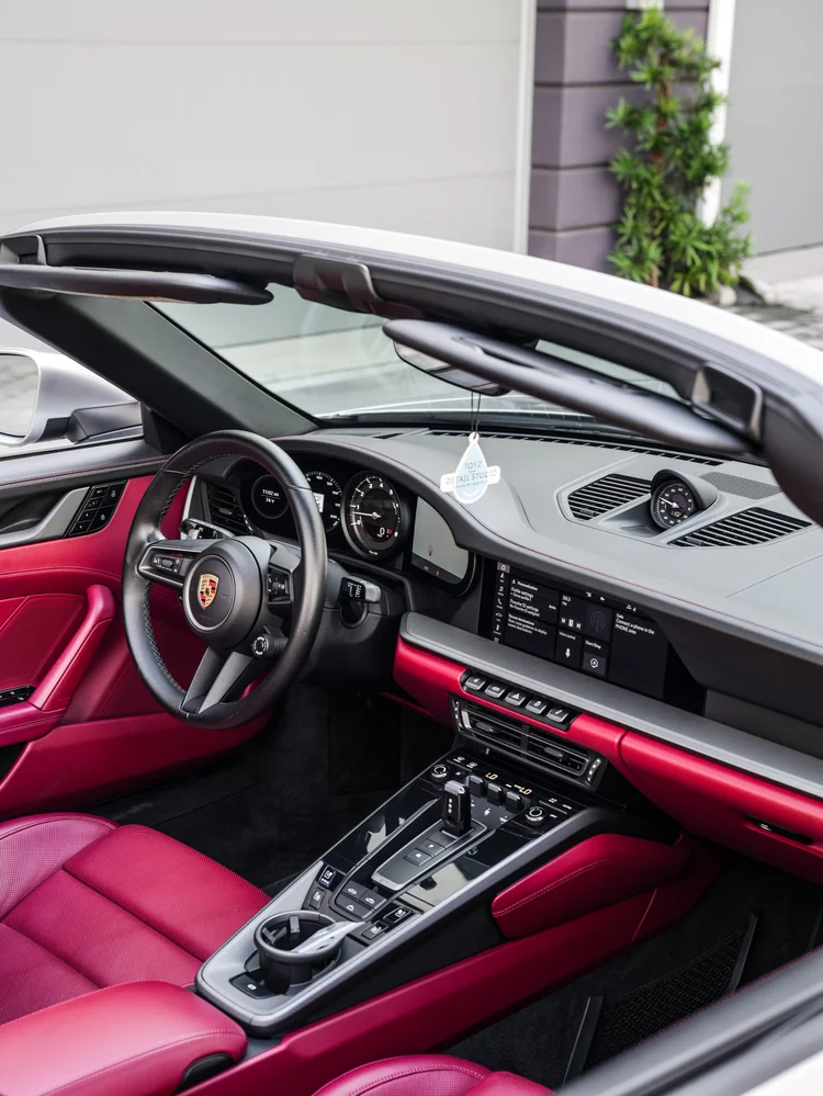 Red and black Porsche cockpit with a crested steering wheel, gauge cluster, dashboard clock, center screen, console, red seat and red door trim.